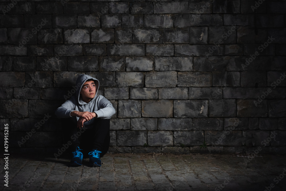 Depressed hispanic teenager sitting against a wall in the darkness. Anxiety and depression in adoloscence concept.