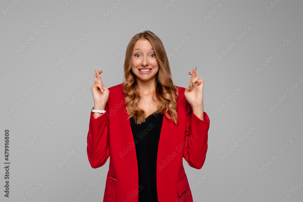 Portrait of happy dreamy young woman with fingers crossed, hopes that she will be lucky, broadly smiling and looking at the camera, standing in red blazer against gray background Portrait of happy dreamy young woman with fingers crossed, hopes that she will be lucky, broadly smiling and looking at the camera, standing in red blazer against gray background