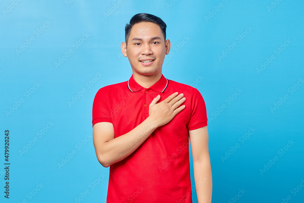 Portrait of handsome asian young man in red shirt holding hands on chest to thank someone and express gratitude isolated on blue background Portrait of handsome asian young man in red shirt holding hands on chest to thank someone and express gratitude isolated on blue background