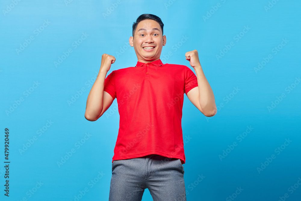 Portrait of excited Asian young man in red shirt celebrating success with cheerful facial expressions on blue background Portrait of excited Asian young man in red shirt celebrating success with cheerful facial expressions on blue background