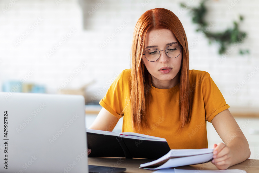 Online learning, portrait of focused beautiful red-haired young female student with laptop and notepad sitting at the table, distance education Online learning, portrait of focused beautiful red-haired young female student with laptop and notepad sitting at the table, distance education