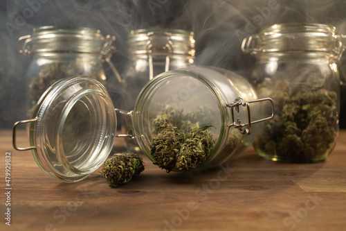 Cannabis buds spilled on the table from a storage glass jar, filled with smoke.
