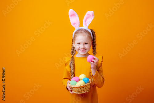 cute little girl with bunny ears holds a basket with Easter eggs on a yellow background