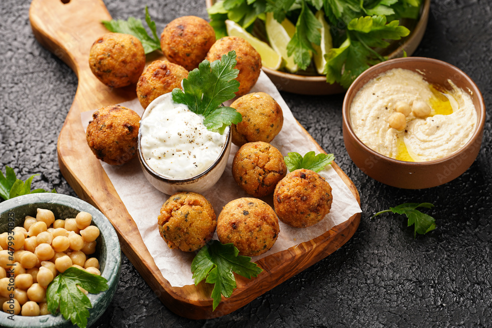 Traditional oriental chickpea deep fried falafel on a wooden board, tzatziki yoghurt sauce, hummus, fresh lime and green cilantro on black surface Traditional oriental chickpea deep fried falafel on a wooden board, tzatziki yoghurt sauce, hummus, fresh lime and green cilantro on black surface
