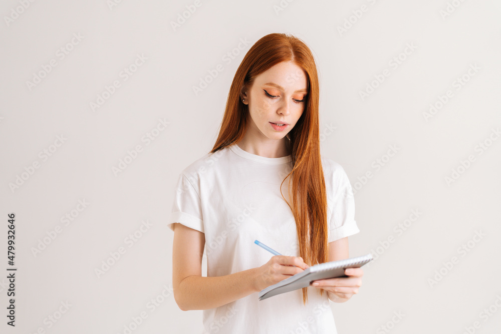Studio portrait of pretty redhead young woman writing in notebook looking down, standing on white isolated background. Confident female student making notes in paper diary. Studio portrait of pretty redhead young woman writing in notebook looking down, standing on white isolated background. Confident female student making notes in paper diary.