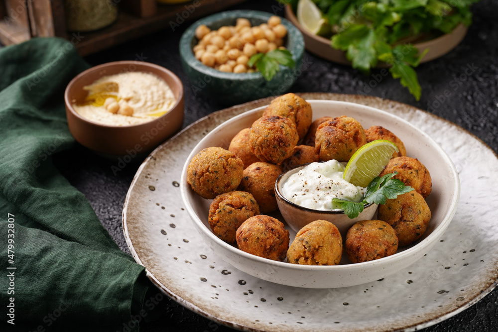 Traditional oriental chickpea deep fried falafel in a deep bowl with tzatziki yoghurt sauce, hummus, fresh lime and green cilantro on black surface Traditional oriental chickpea deep fried falafel in a deep bowl with tzatziki yoghurt sauce, hummus, fresh lime and green cilantro on black surface