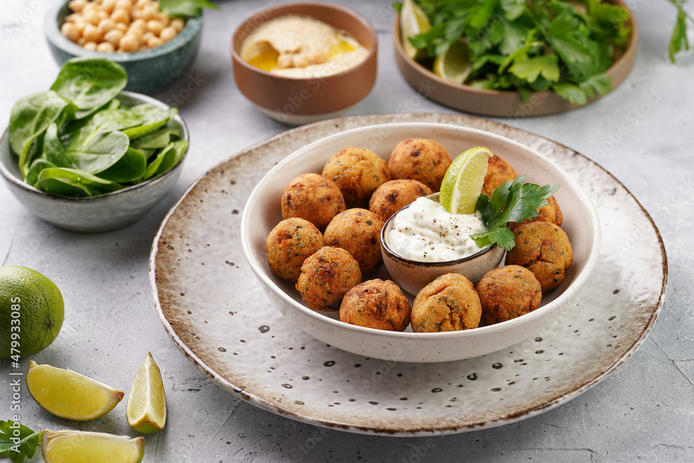 Traditional oriental chickpea deep fried falafel in a deep bowl with tzatziki yoghurt sauce, hummus, fresh lime and green cilantro on light grey concrete surface Traditional oriental chickpea deep fried falafel in a deep bowl with tzatziki yoghurt sauce, hummus, fresh lime and green cilantro on light grey concrete surface