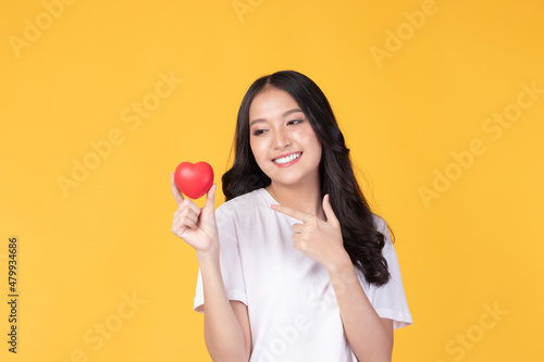 Happy Asian young woman long curly hair holding red heart smiling cute and adorable isolated on yellow background. Love and valentines day.