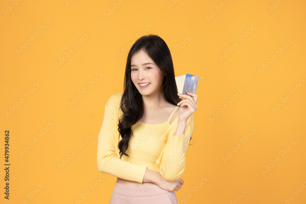 Naklejka premium Cheerful young asian woman in yellow T-shirt holding bank card isolated on yellow background