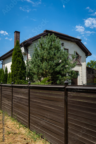 Krasnodar, Russia - Aug 10, 2020: Hedge of evergreen trees behind brown wooden fence with two-story country house. Natural defense against harmful odors and disease-causing bacteria and viruses.