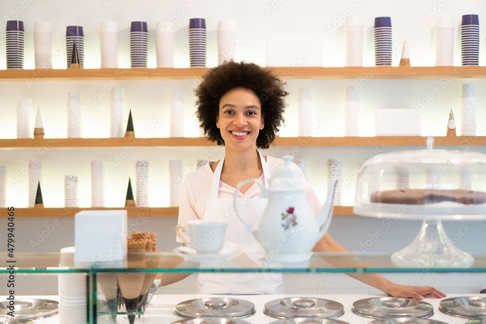 A beautiful mixed-race woman is smiling behind the stand of an ice cream parlor.