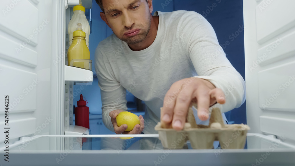 unhappy man puffing cheeks while taking egg and lemon from fridge