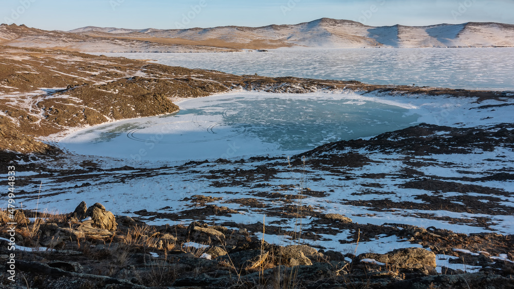 A small frozen heart-shaped lake is surrounded by hills. Round tire tracks are visible on the snow-covered ice. Dry grass on the ground. In the distance - a mountain range against the blue sky. Baikal