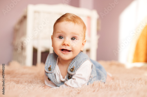 Newborn 5 months old lies on bed on stomach. Baby girl smiles and plays in room. Denim sundress.