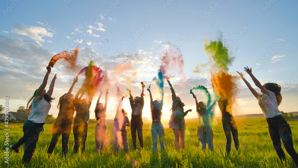 Cheerful girls toss up multi-colored powder at a beautiful sunset. - obrazy, fototapety, plakaty 