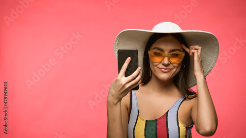 Smiling woman in swimsuit and sunglasses holding smartphone and pointing at head isolated on pink.