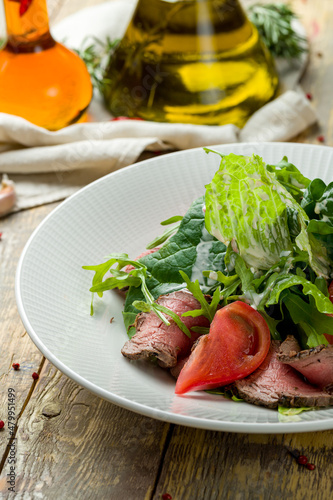 Salad with roast beef, vegetables and greens on white plate on wooden table, vertical