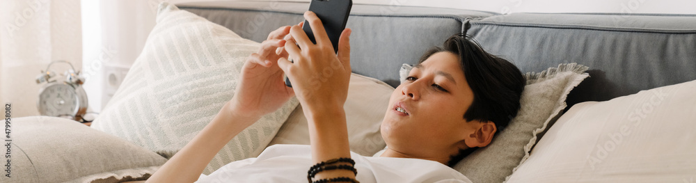 Asian boy using mobile phone while sitting on bed Asian boy using mobile phone while sitting on bed