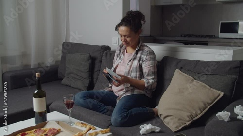 Depressed woman with tear stained face looking at photos, drinking glass of wine, feeling sad, sitting on couch in living room. Unhappy adult female feeling heartbroken after breaking up with lover photo