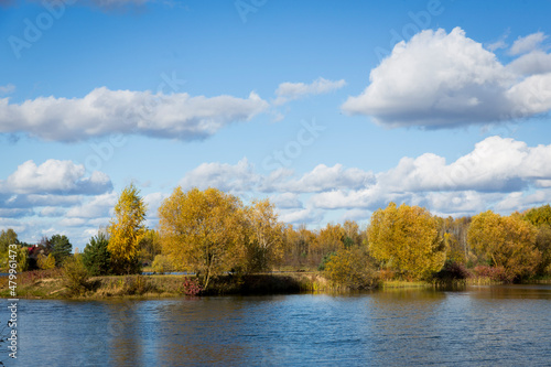 Autumn forest behind the lake. Sky with sun and white clouds. Red-green forest.