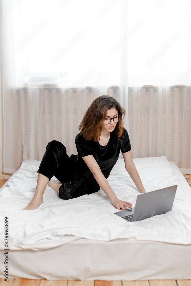 Obraz premium Good morning concept. Beautiful sexy young brunette girl with glasses wearing a black velvet pajama sleepwear on white bed in light cozy bedroom with laptop