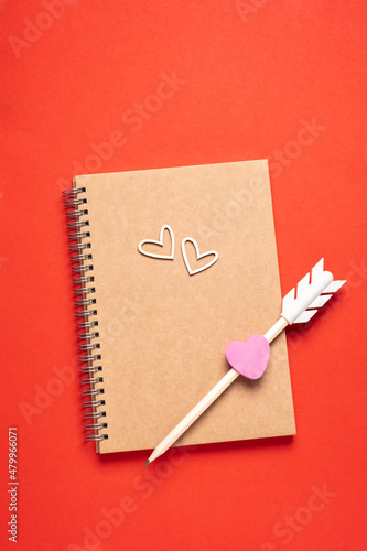 Craft Blank notepad, arrow pensil and wooden hearts on red background. Love message. Valentine's Day and romantic holiday concept. Top view, flat lay with copy space.
