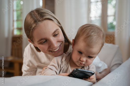 Authentic portrait of happy family using mobile phone watching cartoon, shopping online at home. Mother hugging her cute baby child sitting on sofa © Oleksii
