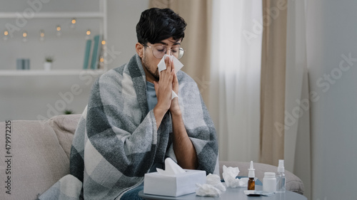 Valokuva Arab bearded sick sad man with glasses sits on couch at home suffers from runny nose flu disease coronavirus pandemic covid epidemic sneezes