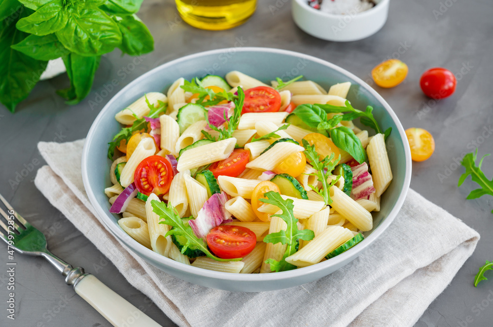 Healthy vegan pasta salad with tomatoes, cucumbers, red onions and arugula in a bowl on a dark gray background. Copy space.