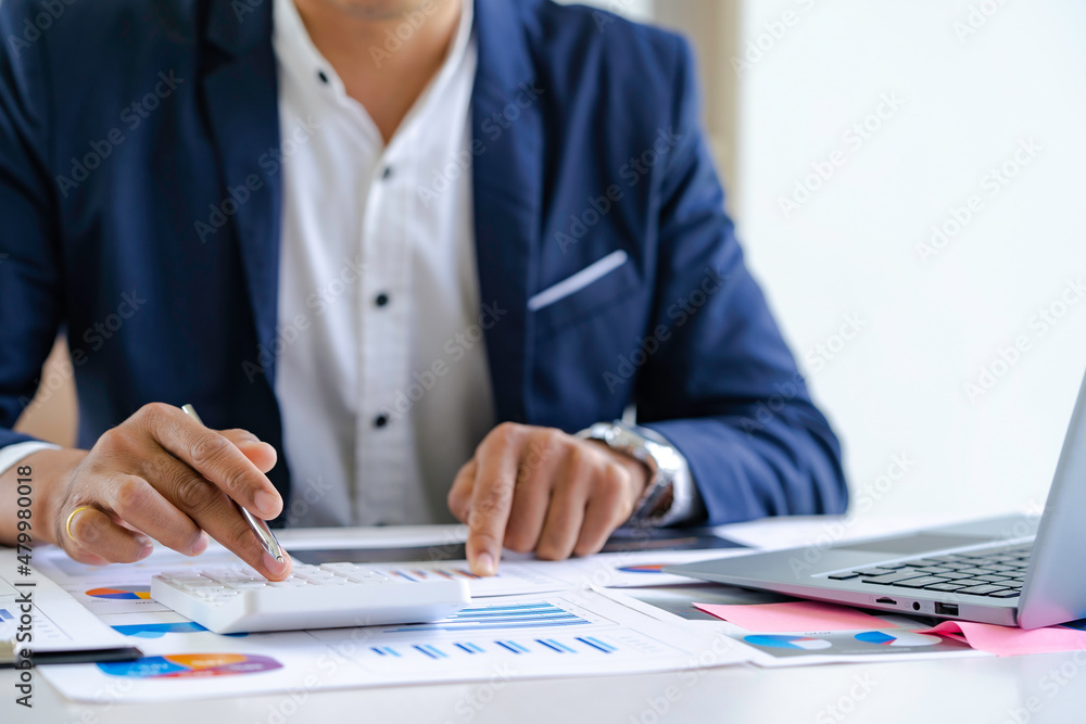 Asian male finance staff is calculating by using a calculator. Investment results to report to his boss at the meeting. On the table in the office, the concept of calculating investment results Asian male finance staff is calculating by using a calculator. Investment results to report to his boss at the meeting. On the table in the office, the concept of calculating investment results