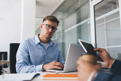 Portrait of Caucasian male employee in classic eyewear for vision protection looking at camera during working process with cropped colleague at table desktop, successful man in optical spectacles