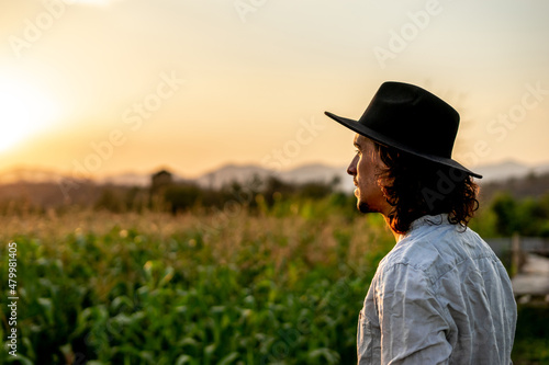 A young hipster farmer looking at the abundance of corn leaves while wearing a hat. An attractive male standing in harmony with the field of nature. Labor, happy hard work, hope, and rich concept