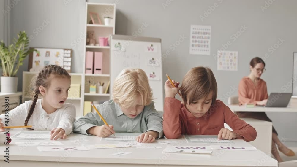 Waist up slowmo shot of three elementary age children sitting at desk in classroom doing test on speech therapy, while their young female teacher working on laptop in background Waist up slowmo shot of three elementary age children sitting at desk in classroom doing test on speech therapy, while their young female teacher working on laptop in background