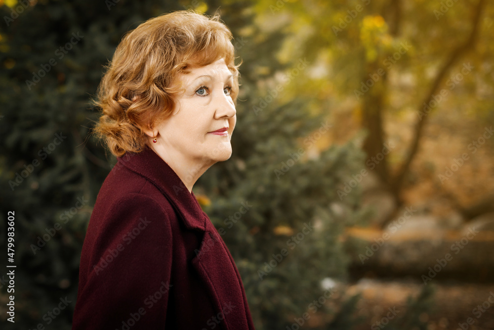 Mature woman in a burgundy coat looks to the side against a background of falling leaves. Mature woman in a burgundy coat looks to the side against a background of falling leaves.