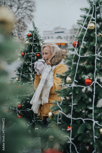 Aged pretty woman with blond hair drinks coffee in the square among the Christmas trees.