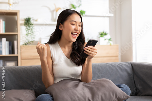 Millennial asian young woman looking mobile phone celebrating with good news or discount voucher for shopping online at home.Happy and cheerful woman looking on cellphone app read message feel excited photo