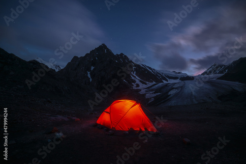 Awesome mountain landscape with vivid orange tent near large glacier tongue under clouds in night starry sky. Tent glow by orange light with view to glacier and mountains silhouettes in starry night. © Daniil