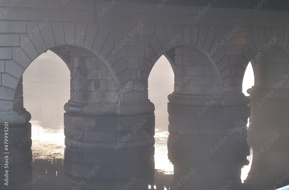 Stone Bridge Supports Reflected in River Waters on Misty Morning 