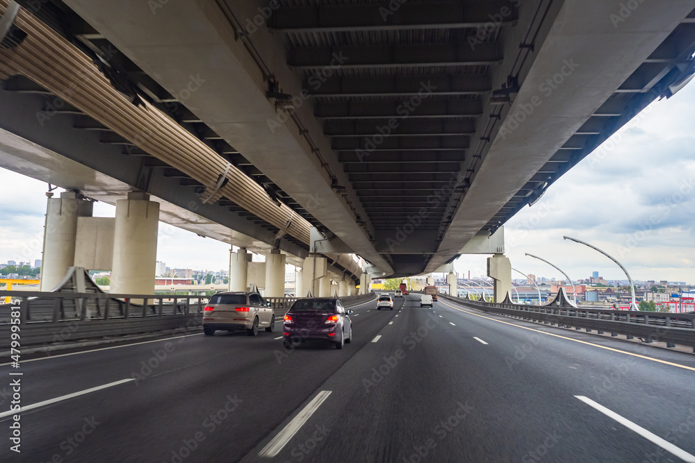 Multilevel road junction. Cars go under railroad, rear view. Multilevel bridge leading to city. Highway. Road Construction Concept. Driveway architecture. Driveway city infrastructure Multilevel road junction. Cars go under railroad, rear view. Multilevel bridge leading to city. Highway. Road Construction Concept. Driveway architecture. Driveway city infrastructure