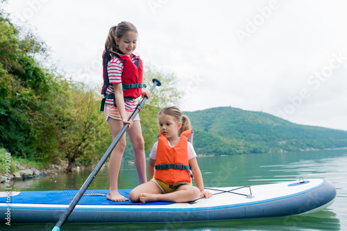 Girls padding on stand up paddle boarding on lake district. Children in swim life vest learning paddleboarding on SUP board. Family active leisure and local getaway with kids concept © tgordievskaya