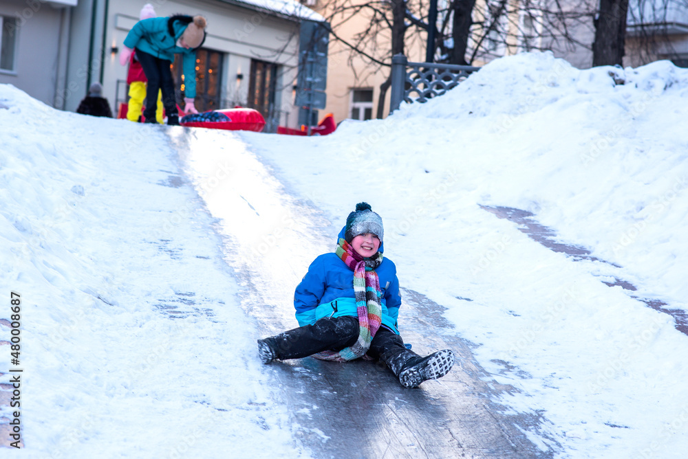 
little boy rides an ice slide in the city. child in blue clothes having fun and resting on winter holidays in the city