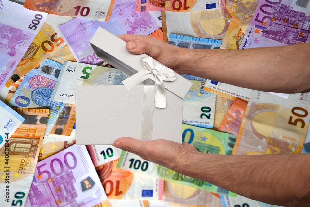 closeup male hands hold white gift, box with satin ribbon against background of paper euro banknotes, concept of cash payments, an expensive gift for holiday, mother's, Valentine's day, birthday