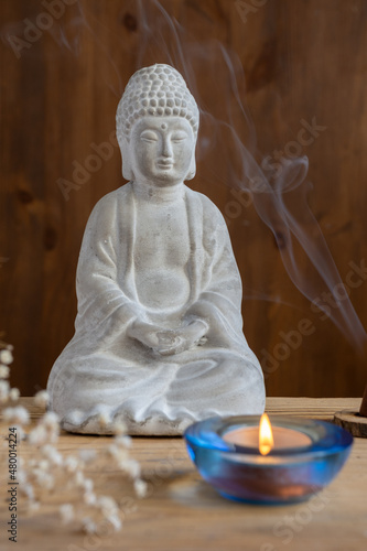 View of sitting white Buddha figure with incense, candle and flowers, selective focus, wooden background, vertical