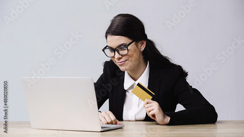 cheerful businesswoman holding credit card near laptop isolated on grey.