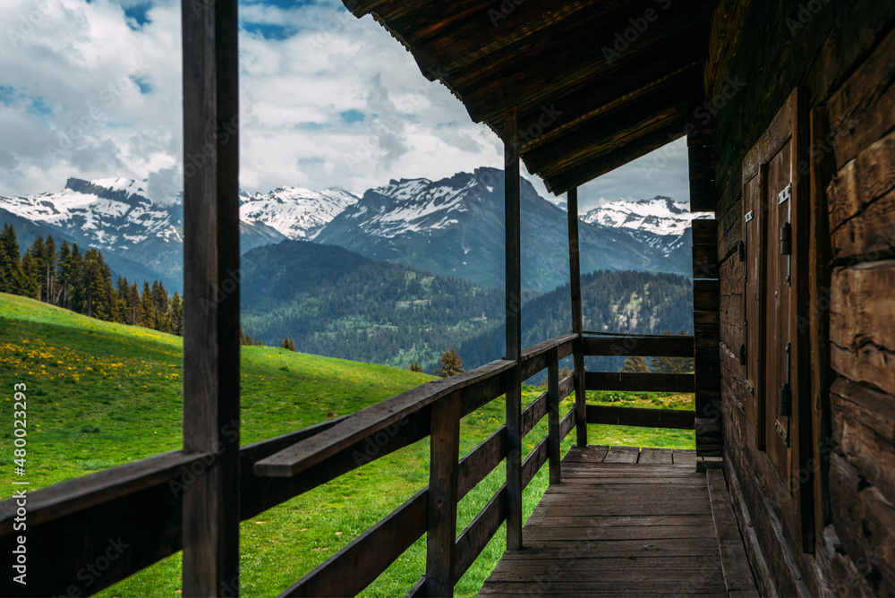 Fototapeta premium Beautiful view of scenic mountain landscape in the Alps with traditional old mountain chalet and fresh green meadows with blue sky and clouds in spring. Part of the facade of a wooden house.
