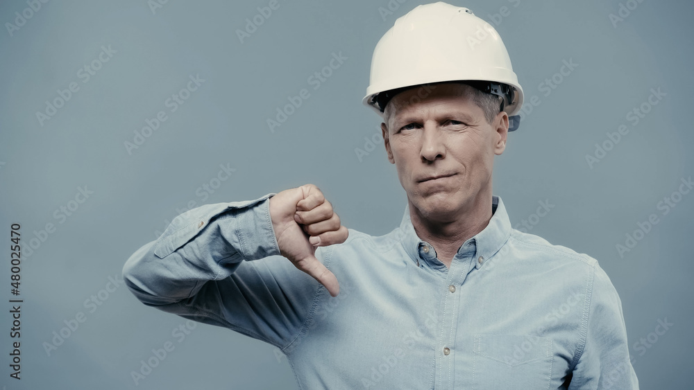 Middle aged engineer in hardhat showing dislike sign isolated on grey.