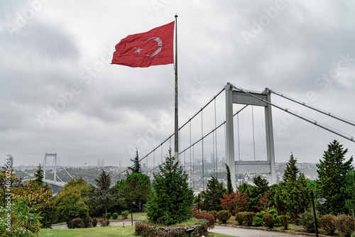 Turkish flag by Fatih Sultan Mehmet Bridge, spanning the Bosphorus strait. The flag of Turkey, officially the Turkish flag is a red flag featuring a white star and crescent. © CanYalicn
