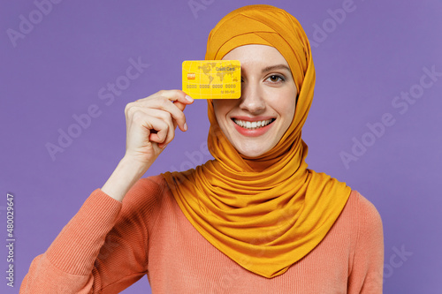 Smiling satisfied jubilant young arabian asian muslim woman in abaya hijab yellow clothes cover close hiding eye with credit bank card isolated on plain pastel light violet background studio portrait. photo