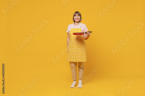 Full size elderly fun happy smiling housekeeper housewife woman 50s in orange apron hold empty frying pan cook isolated plain on yellow background studio portrait. People household lifestyle concept.