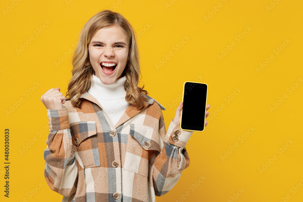 Little smiling surprised happy blonde kid girl 13-14 years in checkered shirt hold in hand use mobile cell phone with blank screen workspace area do winner gesture isolated on plain yellow background. Little smiling surprised happy blonde kid girl 13-14 years in checkered shirt hold in hand use mobile cell phone with blank screen workspace area do winner gesture isolated on plain yellow background.